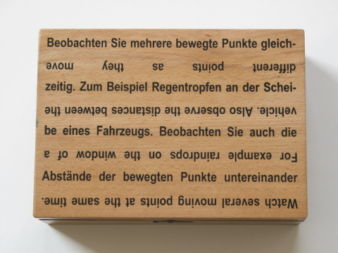 Holzkasten mit folgender Aufschrift: Beobachten Sie mehrere bewegte Punkte gleichzeitig. Zum Beispiel Regentropfen an der Scheibe eines Fahrzeugs. Beobachten Sie auch die Abstände der bewegten Punkte untereinander. Watch several moving points at the same time. For example raindrops on the window of a vehicle. Also observe the distances between the diffrent points as they move.