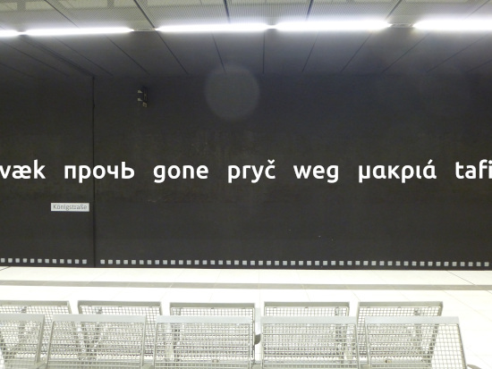 Auf der Wand einer unterirdischen Bahnstation steht in weißer Schrift auf schwarzem Grund der Begriff weg in verschiedenen Sprachen.